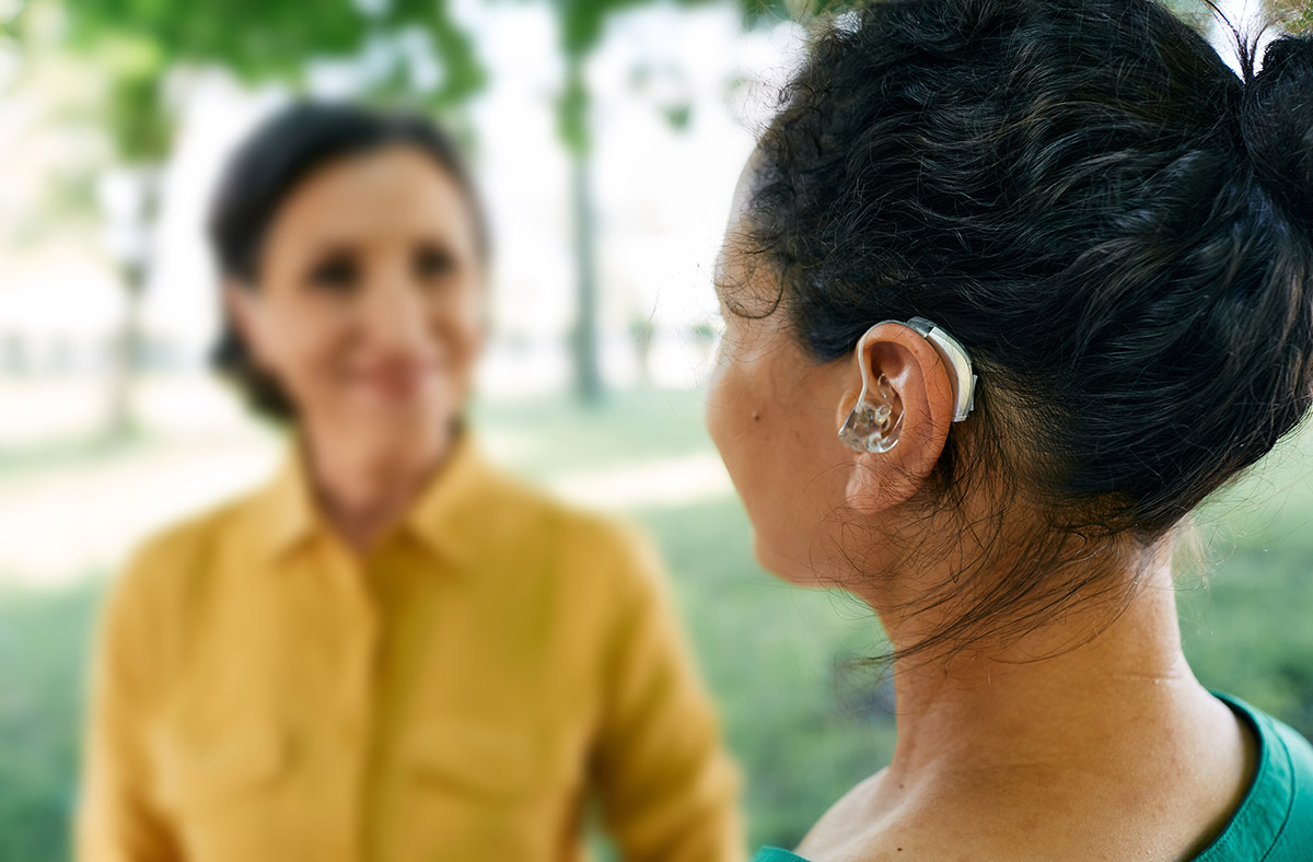 Back view of the head of a woman wearing a hearing aid, she is having a conversation with a smiling woman