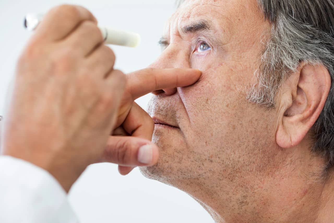 Image of an older man's going through a medical examination of his left eye, only the doctor's hands are visible, one is holding the man's cheek and the other is pointing a light to the patient's eye