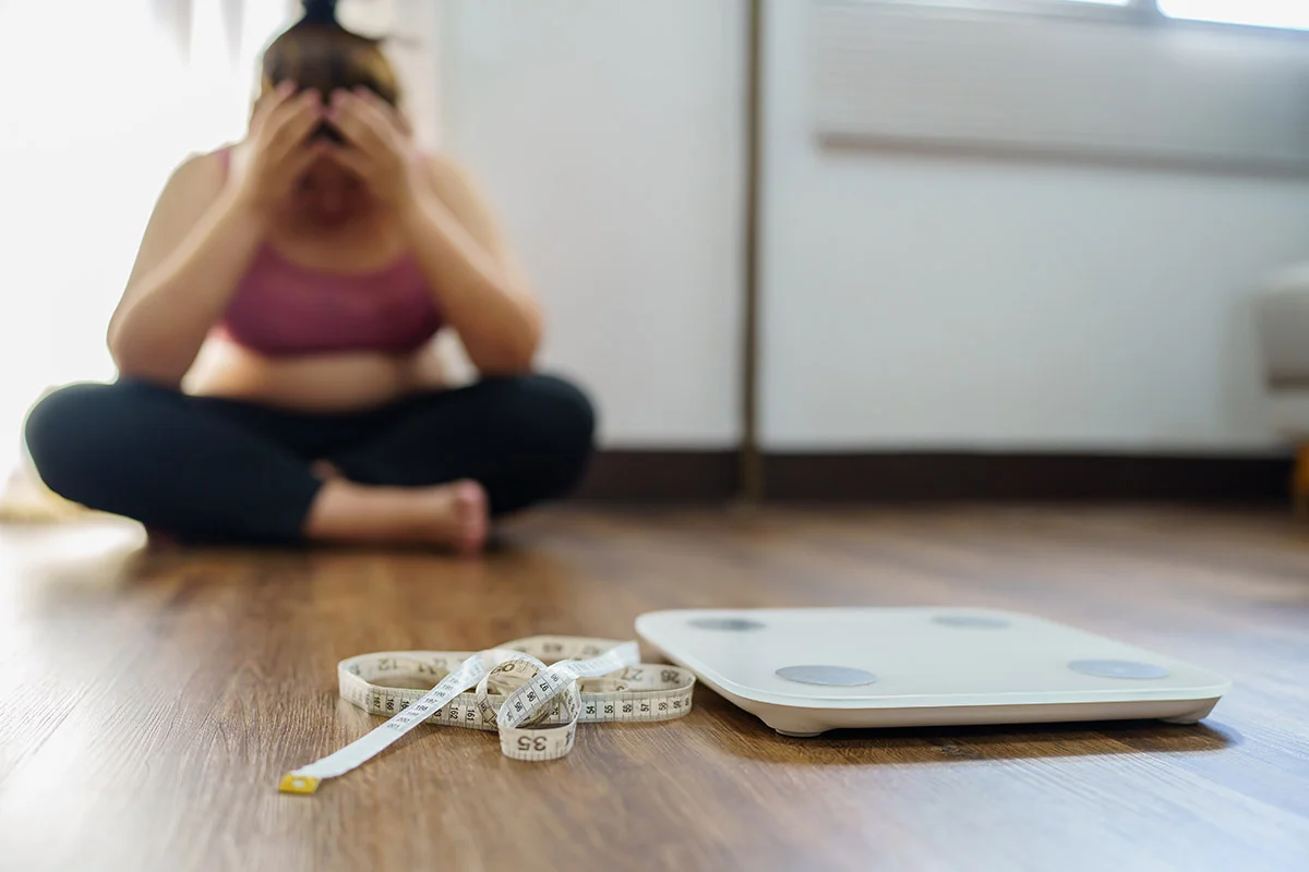 disheartened woman sitting on the kitchen floor with head in hands with a weight scale in the foreground